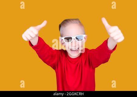 un enfant gai dans des vêtements et des lunettes rouges montre les pouces sur un fond jaune Banque D'Images