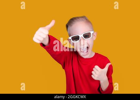 un enfant gai dans des vêtements et des lunettes rouges montre les pouces sur un fond jaune Banque D'Images