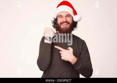 Un jeune homme à barbe heureux portant un chapeau de Noël pointe vers sa montre à bracelet sur fond blanc. Banque D'Images