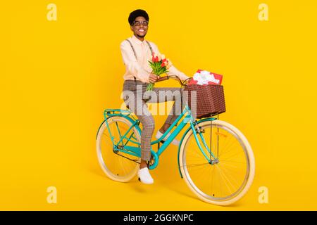 Photo de gai bien entretenu gars conduire vélo tenir fleurs porter des bretelles chemise isolé couleur jaune fond Banque D'Images