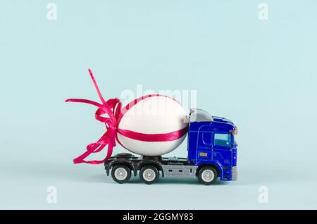 Le tracteur jouet transporte un œuf de poulet. Camion en plastique bleu et œuf blanc avec ruban rouge. Arrière-plan bleu. Camionnage. Mise au point sélective. Banque D'Images