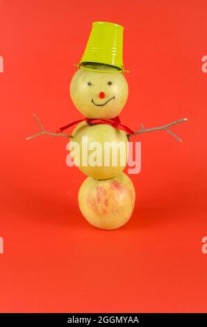 Un bonhomme de neige aux pommes debout sur un fond rouge. Bonhomme de neige créatif de trois fruits mûrs avec un seau sur sa tête. Banque D'Images