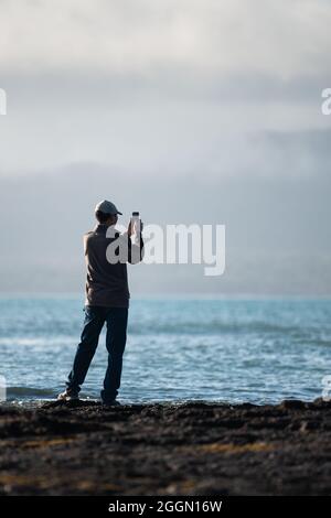 Homme prenant des photos à l'aide d'un téléphone portable sur la plage de Milford. Arrière-plan l'île Rangitoto dans la brume. Format vertical. Banque D'Images