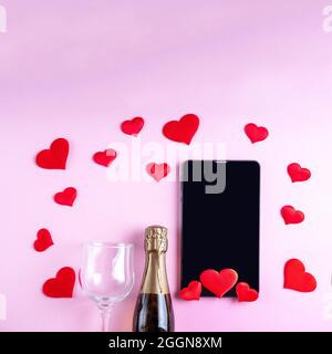 Félicitations en ligne pour la Saint-Valentin. Bouteille de champagne, verre de vin, tablette noire ou écran de téléphone sur fond rose, décoré de coeurs rouges, s Banque D'Images