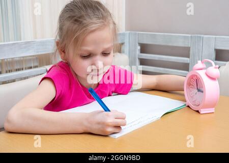 Une petite fille s'assoit à la table et écrit ses devoirs dans un bloc-notes. Temps d'étude. L'enfant étudie à la maison. Concept d'apprentissage à domicile Banque D'Images