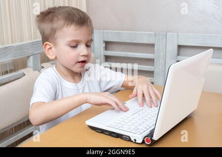 Un adorable petit garçon est assis à une table et tape ses devoirs sur un ordinateur portable à la maison. Concept d'apprentissage, apprentissage à distance, auto-éducation, vidéo conferenci Banque D'Images