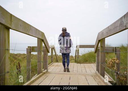 Vue arrière d'une jeune femme marchant sur une promenade en direction de la plage et de la mer en face d'elle lors d'une journée couverte au pays de Galles Banque D'Images