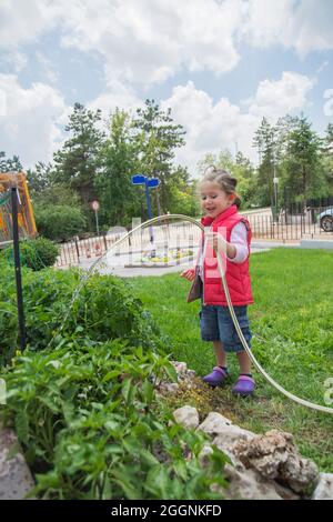 La petite fille arrose les plants de tomates et de poivrons dans leur jardin avec un tuyau de jardin. Sélectif Focus. Banque D'Images