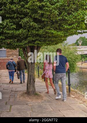 Un chemin avec des arbres et des bancs longe une rivière. Deux couples, jeunes et vieux, marchent le long du chemin avec un pont en arrière-plan. Banque D'Images