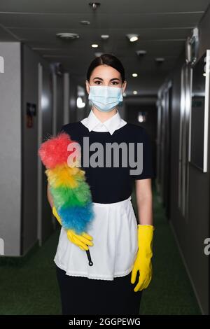 femme de ménage dans un masque médical et des gants en caoutchouc tenant une brosse à poussière dans le couloir de l'hôtel Banque D'Images