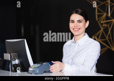 bonne réceptionniste souriant près du lecteur de carte de crédit sur le comptoir Banque D'Images