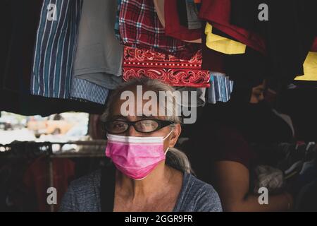 Portrait d'une femme mature fournisseur de vêtements avec un masque le jour du marché. Belmopan, Belize. Banque D'Images