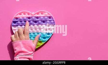 Coeur en forme de bouton il antistress jouet avec la main femelle. Un simple violon sensoriel aux couleurs des LGBT. Couché à plat sur du papier rose avec espace de copie Banque D'Images