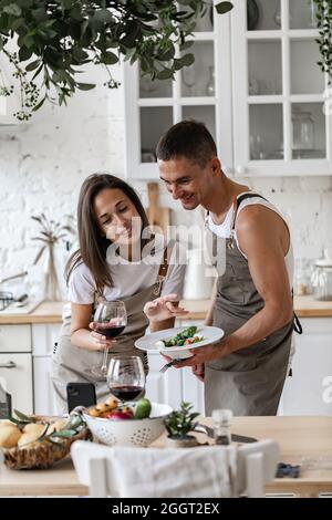 Couple heureux de passer un appel vidéo avec un smartphone tout en préparant le dîner dans la cuisine à la maison les gens gais de Caucasiens agités leurs mains, souriant Banque D'Images