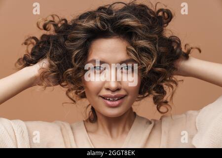 Jeune femme hispanique mauriquement souriante et touchant ses cheveux isolés sur fond beige Banque D'Images