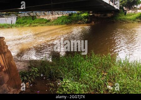 Une rivière d'eau brunâtre coule sous un pont. Sur la gauche et la droite de la rive de la rivière, l'herbe pousse verte, et un peu de détritus peut être vu. Banque D'Images