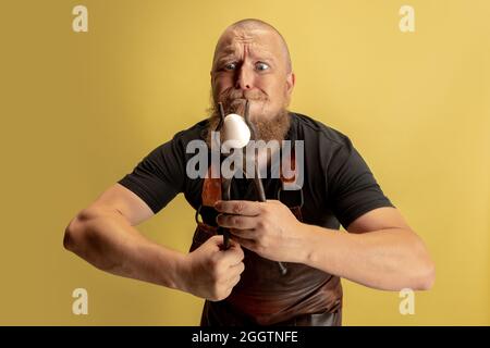 Portrait comique de chauve barbu musclé, forgeron en tablier de cuir ou uniforme isolé sur fond jaune de studio. Concept de travail, rétro Banque D'Images