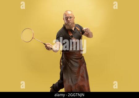 Portrait comique de chauve barbu musclé, forgeron en tablier de cuir ou uniforme isolé sur fond jaune de studio. Concept de travail, rétro Banque D'Images