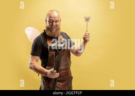 Portrait comique de chauve barbu musclé, forgeron en tablier de cuir ou uniforme isolé sur fond jaune de studio. Concept de travail, rétro Banque D'Images