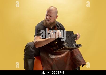 Portrait comique de chauve barbu musclé, forgeron en tablier de cuir ou uniforme isolé sur fond jaune de studio. Concept de travail, rétro Banque D'Images