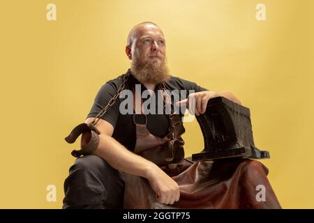 Portrait comique de chauve barbu musclé, forgeron en tablier de cuir ou uniforme isolé sur fond jaune de studio. Concept de travail, rétro Banque D'Images