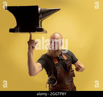 Portrait comique de chauve barbu musclé, forgeron en tablier de cuir ou uniforme isolé sur fond jaune de studio. Concept de travail, rétro Banque D'Images