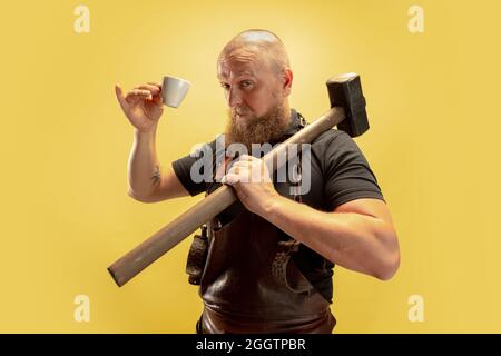 Portrait comique de chauve barbu musclé, forgeron en tablier de cuir ou uniforme isolé sur fond jaune de studio. Concept de travail, rétro Banque D'Images
