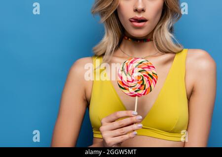 coupe courte de la jeune femme en haut de bikini jaune qui dépasse la langue et tient la douce lollipop sur le bleu Banque D'Images