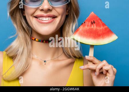 vue courte de la jeune femme heureuse en lunettes de soleil tenant le bâton de popsicle avec la pastèque isolée sur le bleu Banque D'Images
