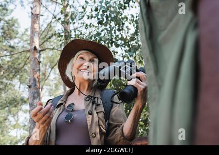 Vue à angle bas d'un voyageur senior heureux tenant des jumelles près d'un mari afro-américain flou dans la forêt Banque D'Images