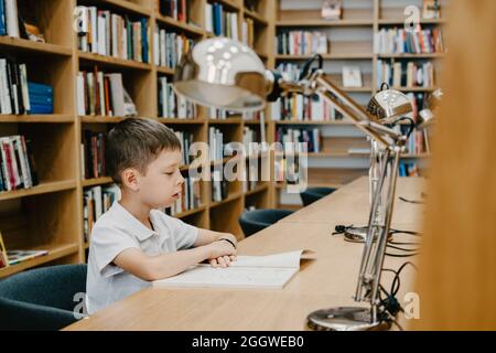 Un écolier assis sur une table et lisant un livre à la bibliothèque à l'école. Préparer pour les devoirs. Un bon étudiant. Le garçon aime lire. Espace libre au niveau du sch Banque D'Images