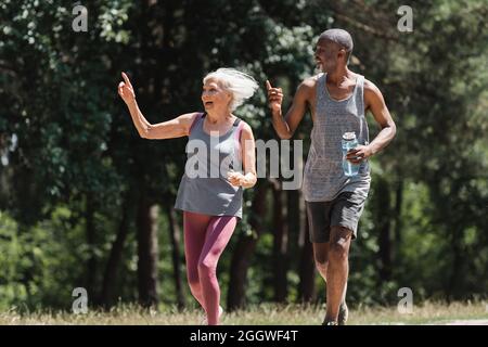 Couple multiethnique heureux avec bouteille de sport pointant avec les doigts pendant la course dans le parc Banque D'Images