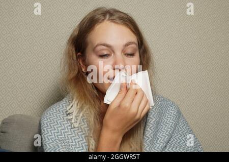 La femme éternue dans un mouchoir blanc. Jeune femme blonde enveloppée dans une couverture chaude, éternuant, a la grippe. Banque D'Images