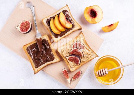 Beurre d'arachide, tartiner au chocolat avec des fruits sur un toast sur une planche à découper sur fond blanc, petit déjeuner sucré sain, gros plan. Banque D'Images