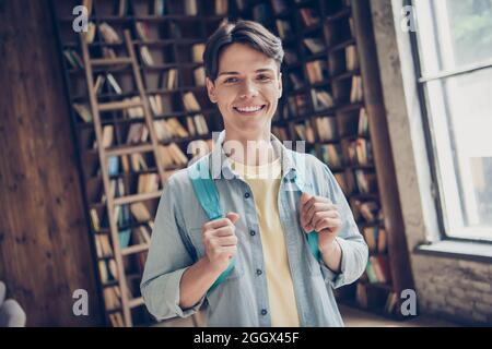 Photo de gai souriant bonne humeur université étudiant porter sac à dos Jean denim chemise détente à l'intérieur dans la bibliothèque Banque D'Images