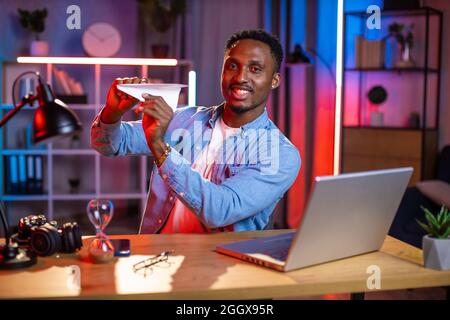 Homme afro-américain en chemise denim jouant avec un avion en papier tout en utilisant un ordinateur portable pendant la soirée. Jeune travailleur indépendant assis à son bureau et distrait du travail à la maison. Banque D'Images