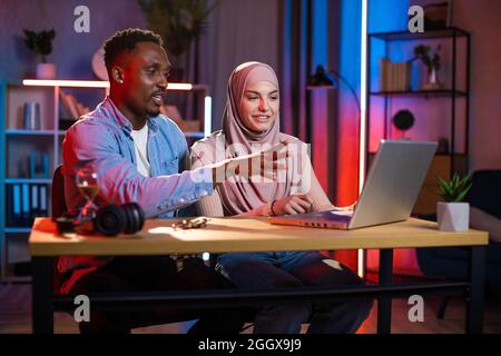 Homme africain et femme arabe assis ensemble au bureau à domicile et regardant sur l'écran d'ordinateur portable. Collègues multiraciaux discutant d'un projet commun pendant la soirée. Concept de coopération et de travail d'équipe. Banque D'Images