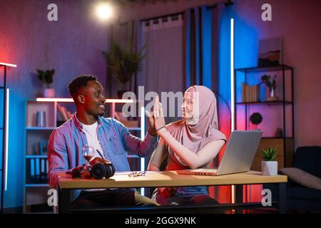 Homme africain donnant cinq hauts à la femme dans hijab tout en s'asseyant ensemble au bureau avec un ordinateur portable moderne. Coopération réussie des partenaires multiraciaux. Concept de personnes, d'entreprises et de technologie. Banque D'Images