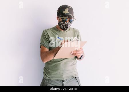 Militaire avec cagoule de camouflage, lunettes de soleil et masque de camouflage, écrivant un rapport dans un dossier, sur un mur blanc Banque D'Images