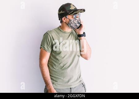 Militaire vêtu d'un chapeau de camouflage, de lunettes de soleil et d'un masque de camouflage, parlant sur le téléphone portable, sur un mur blanc Banque D'Images