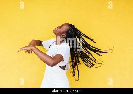 Vue latérale d'une jeune femme heureuse avec de longues tresses dansant au mur jaune à l'extérieur Banque D'Images