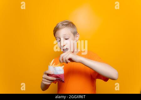 Un garçon dans un T-shirt orange tient un sac de pommes de terre frites en face de lui et en prend un morceau Banque D'Images