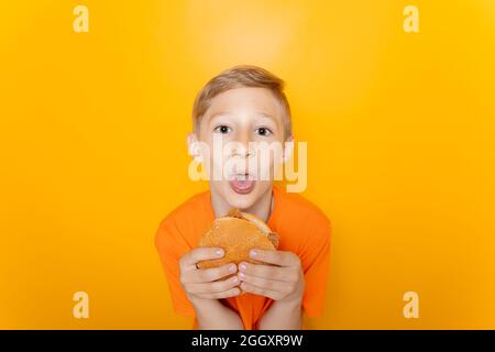 Un garçon dans un T-shirt orange tient un hamburger devant lui et colle sa langue à l'appareil photo Banque D'Images