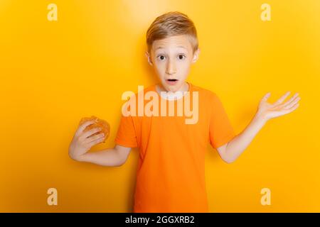 Un garçon dans une chemise T orange tient un hamburger dans une main, déploie ses bras sur les côtés et montre un geste de brewilderment Banque D'Images