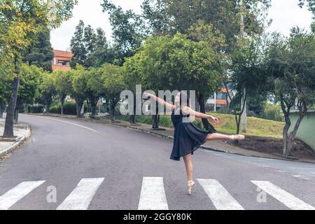 Danseuse de ballet adolescente avec robe noire debout sur un orteil dans une pose de ballet sur un zébré traversant une rue de ville Banque D'Images