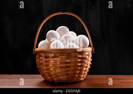 Un panier en osier composé d'œufs blancs fraîchement collectés sur une table en bois et encadré d'un arrière-plan subtil noir marbré. Banque D'Images