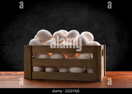 Une petite caisse en bois marron d'œufs blancs fraîchement collectés sur une table en teck et encadrée d'un arrière-plan subtil noir marbré. Banque D'Images