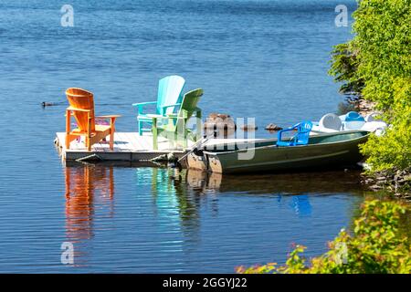 Trois chaises Adirondack colorées sur un quai flottant entouré d'eau encore bleue. Les chaises sont de couleur orange, verte et bleue. Banque D'Images