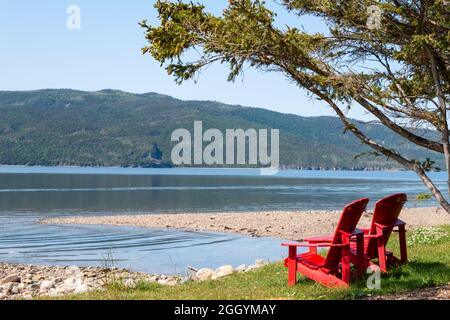 Deux chaises Adirondack rouge vif et vide sur le bord d'un pré vert herbacé surplombant l'océan bleu avec des montagnes couvertes d'arbres. Banque D'Images
