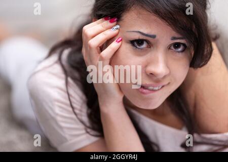 Une jeune femme inquiète et attentiste se mordant sur la lèvre, donnant à l'appareil photo un aspect anxieux tout en se relaxant sur un canapé à la maison dans un portrait rapproché Banque D'Images